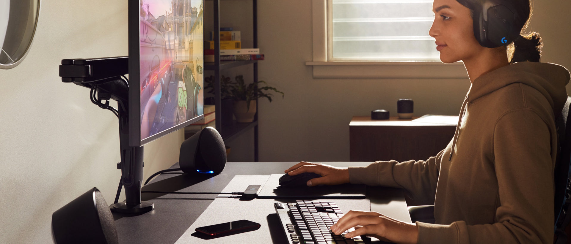 Una donna con una felpa indossa degli auricolari e ha una mano sul mouse e l’altra sulla tastiera, mentre guarda un monitor collocato su un braccio porta monitor Ollin.