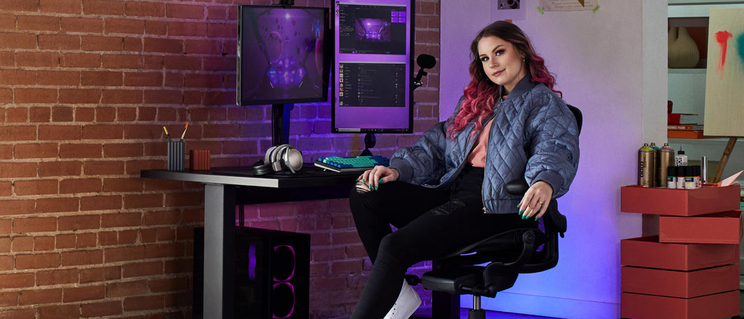 Una donna dai capelli rosa si rilassa sulla seduta per gaming Aeron mentre occupa la sua postazione di gioco, circondata da grandi monitor e una scrivania sit-stand.