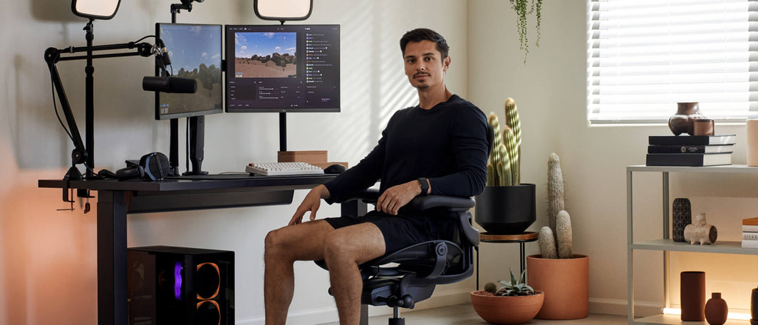 Gamer vestito completamente di nero su una seduta per gaming Aeron, davanti alla sua spaziosa configurazione di gioco, con monitor, un microfono e piante in vaso.